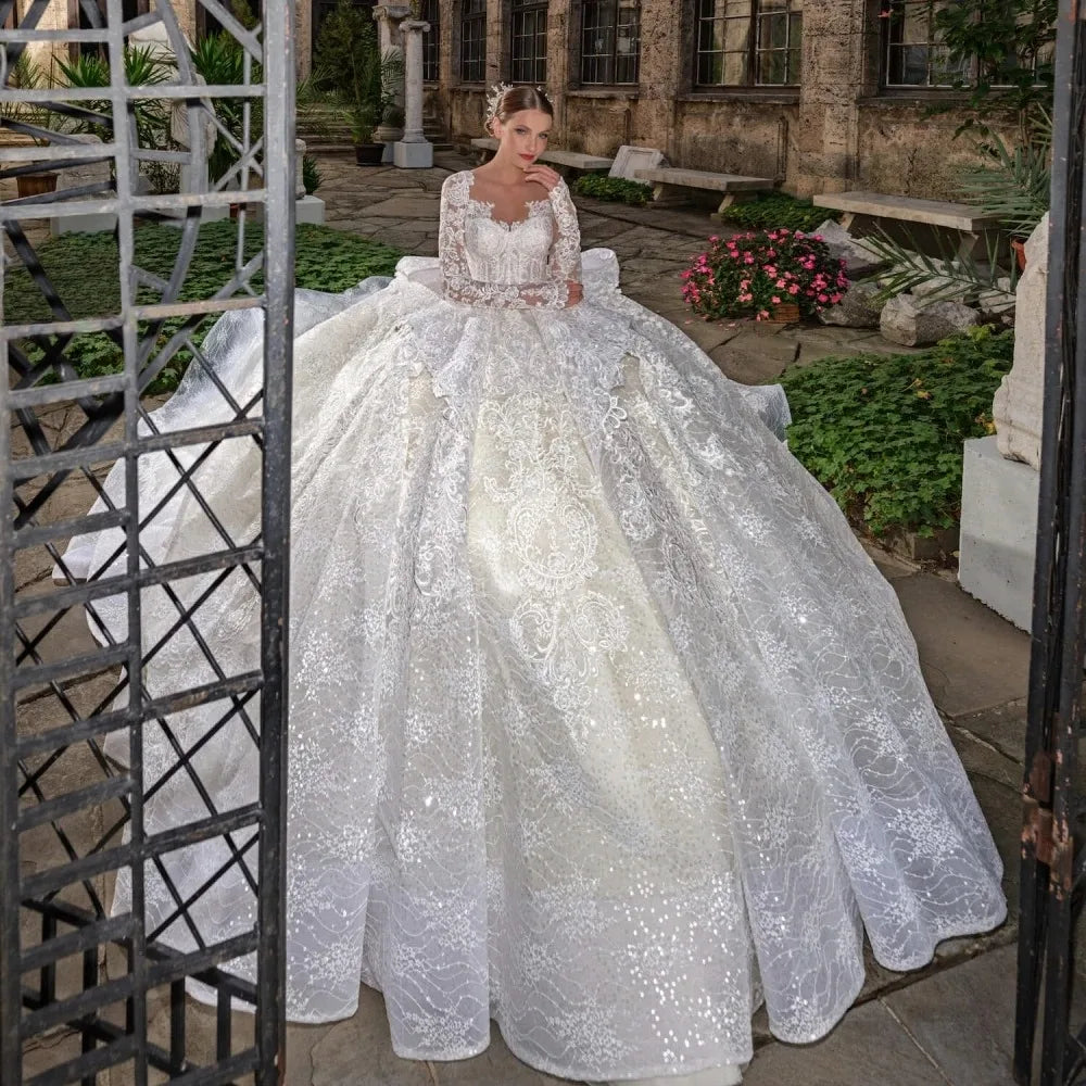 Woman in a white lace wedding dress standing outdoors with greenery and a building in the background.
