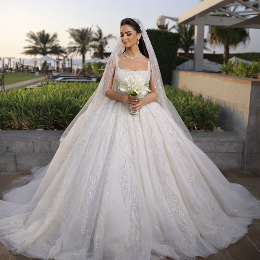 Woman in a white wedding dress holding flowers outdoors with greenery and a building in the background